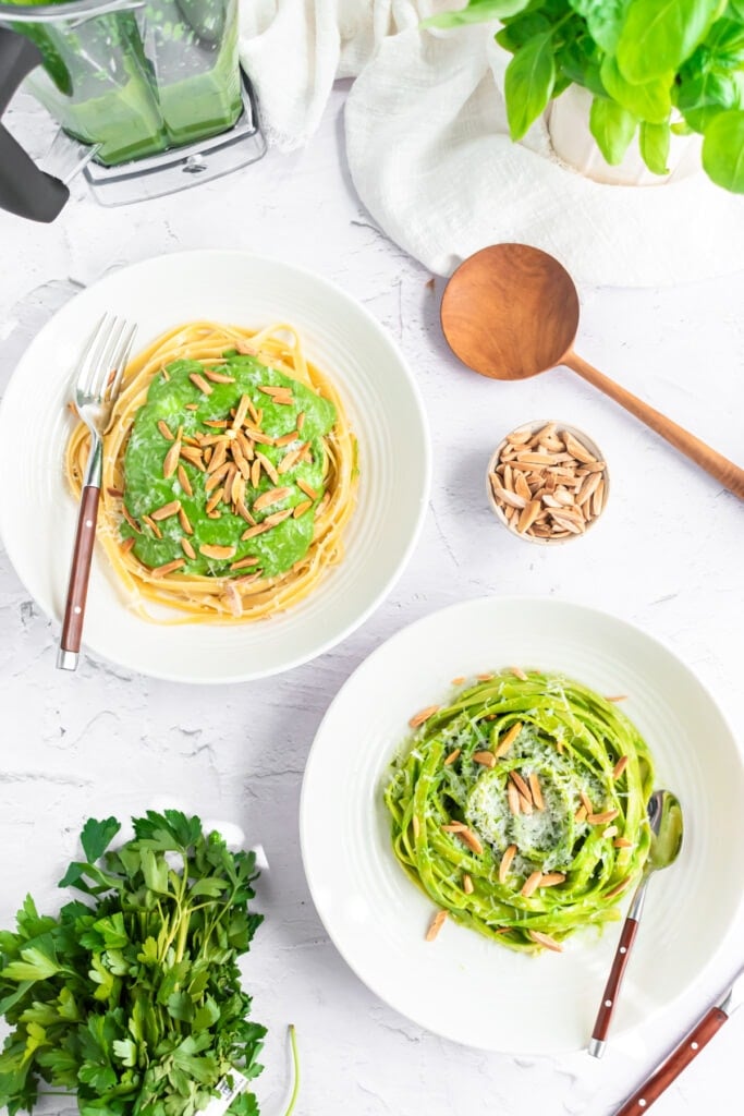Two white bowls of green spaghetti sauce. One is topped with green sauce and slivered almonds, the other pasta has been coated with the green sauce. The bowls sit on a white surface. Nearby are fresh parsley, a bowl of almonds, a wooden spoon, and a blender jar filled with the vibrant sauce.