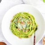 an overhead shot of fettuccini pasta coated in green spaghetti sauce in the center of a shallow white bowl topped with parmesan and toasted almond slivers