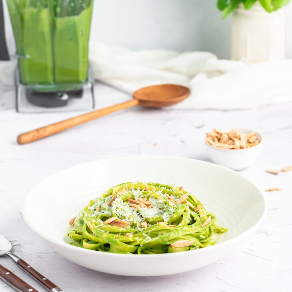 A white bowl of green spaghetti fettuccine topped with grated cheese and slivered almonds sits on a white surface, with a small bowl of almonds, wooden spoon, and a blender of green sauce in the background.