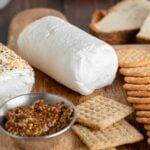 a log of coat cheese in the middle of a wooden board. a slice has been cut off the front of the log. small bowls of capers and mustard are to the left. a stack of crackers is to the right.