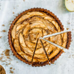 an overhead shot of a pear and gruyere tart with three slices cut out of the bottom right section of it. two halves of a pear are visible in the top right corner and a small bowl of brown sugar in the bottom left. two forks lie next to the tart in the top left.