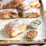 a close up of a ciabatta loaf on a sheet pan with a bulb of roasted garlic and pinch bowl of fennel seeds next to it. more ciabatta loaves are in a pile behind it.