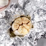 a bulb of garlic with the top sliced off covered in a drizzle of olive oil and sprinkle of salt and pepper sits in the middle of a crumpled sheet of aluminum foil