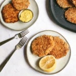 an overhead shot of two tuna cakes on a small grey plate with half of a whole lemon. two additional plates with more tuna cakes are on the table above it. one of them has a small bowl of aioli on it.