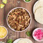 an overhead shot of shredded pork carnitas in a serving bowl surrounded by the other ingredients for making tacos.