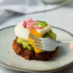 a fork pierces through the side of a poached egg perched on top of guacamole on a small potato pancake