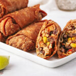 an overhead shot of four egg rolls sliced on a diagonal lined up next to each other on a small grey plate with two lime wedges. next to the plate is half a lime and a small bowl of dipping sauce.