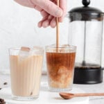 two glasses of cold brew coffee with milk. one glass is being stirred. an empty french press is behind them.