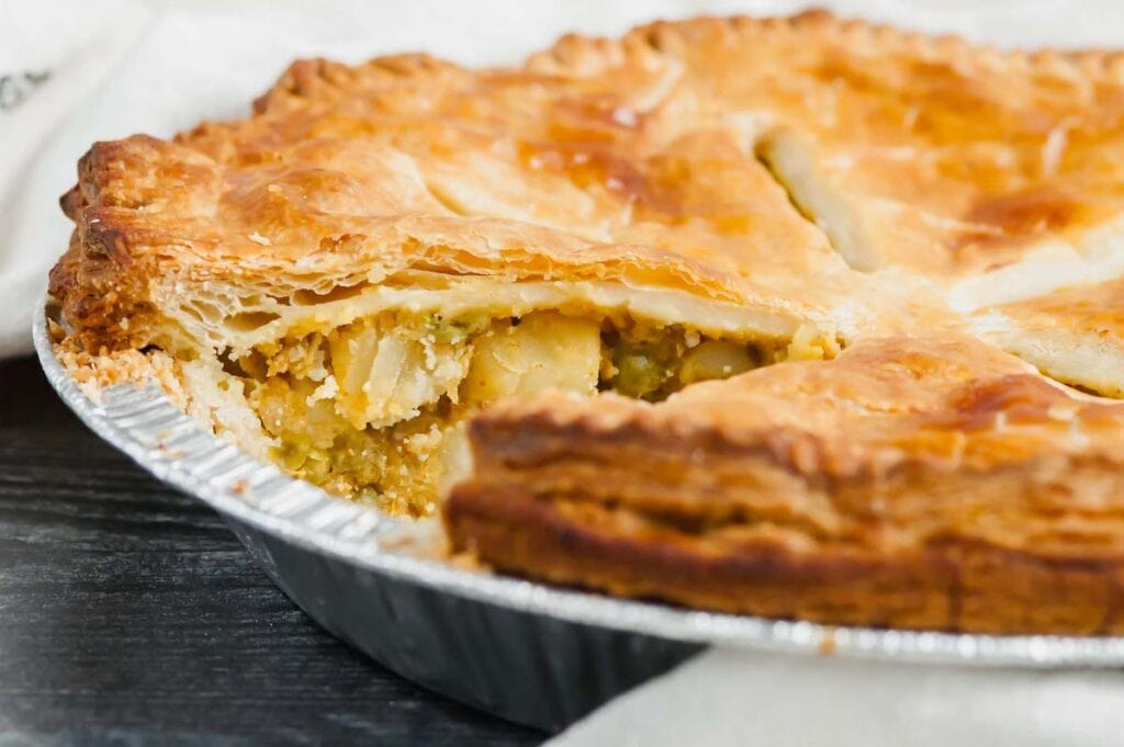 A close-up of a golden-brown samosa pot pie in a foil pan with a slice removed, revealing a filling of potatoes and peas. The crust looks flaky and crisp.