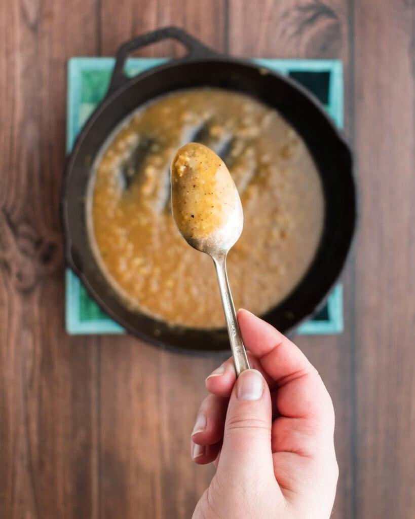 A hand holds a spoonful of brown gravy above a cast iron skillet filled with more of the same gravy, set on a wooden surface—perfect for serving alongside juicy cast iron pork tenderloin.