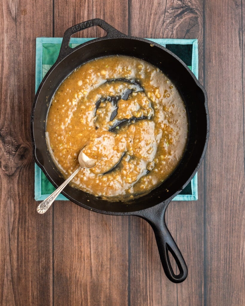 A cast iron pork tenderloin simmers in a skillet filled with thick, brown sauce, set atop a blue cloth on a wooden surface. A metal spoon rests in the skillet, partially coated with the savory sauce.