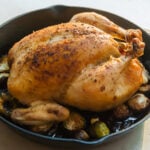a whole roast chicken sits on a bed of brussels sprouts in a cast iron pan on a metal counter