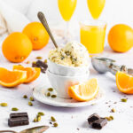 a scoop of orange cardamom ice cream in a small white bowl surrounded by orange slices, cardamom pods, and chocolate squares