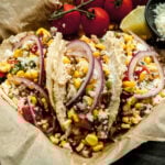 a close up of 3 tacos in a parchment lined sheet pan with a small bowl of cotija cheese, some lime wedges, and whole cherry tomatoes