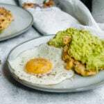 two corn fritters topped with mashed avocado on a small plate with a fried egg. in the backgroud another plate with corn fritters on it is visible.