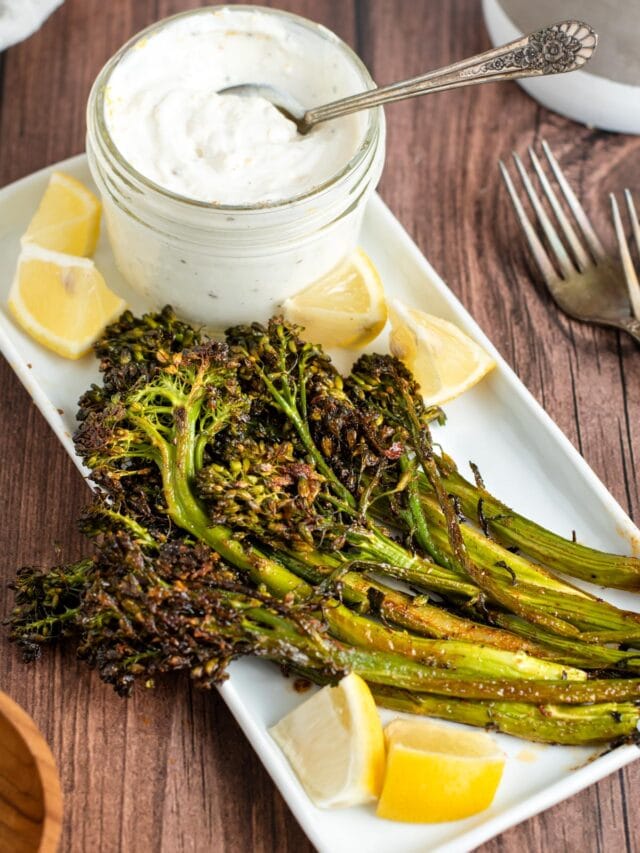 roasted broccolini on an rectangle platter with a small jar of lemon yogurt dipping sauce and lemon wedges