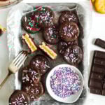 an overhead shot of a tray of patty cakes. one has been cut in half with the insides facing up showing the layers of peanut butter and cracker inside. a small bowl of blue purple pink and white sprinkles is onn the tray. crackers and chocolate are to the right of the tray and a fork with its tines dripping chocolate rests on the edge.