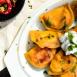 an overhead shot of an oval platter of tikka masala pierogis topped with a dollop of greek yogurt sauce and garnished with cilantro. a small bowl with two red chilies and cardamom pods sits to the left.