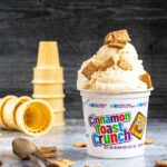 scoops of ice cream piled high in a small plastic container for cinnamon toast crunch topped with pieces of the cereal. some ice cream cones are stacked up in the background.