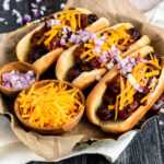 three chili hot dogs in a dish with two small bowls filled with shredded cheddar and minced red onion