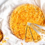 an overhead shot of a broccoli quiche. the bottom quarter of the quiche has been cut into 3 wedges, one of which is turned on its side. to the left is a small bowl of shredded cheddar cheese.