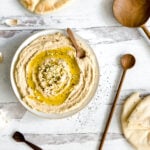 an overhead shot of a bowl of hummus drizzled with olive oil and minced garlic on a weathered white wooden countertop. three wooden spoons, a torn pita bread, and some garlic cloves are on the table next to the hummus.
