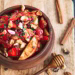 an overhead shot of a wooden bowl filled with fruit salad. the bowl sits on a wooden board next to a honey wand and two cinnamon sticks.