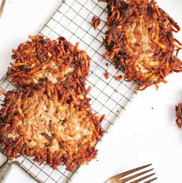 an overhead shot of potato pancakes on a narrow wire cooling rack. two are stacked on top of each other, one sits alone. another is cropped just out of frame.