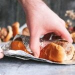 two hands wrapping a sage sausage in a hot dog bun in a sheet of aluminum foil