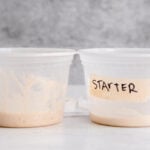 two 16 oz deli containers sit side by side. the one on the right has about half an inch of mature sourdough starter in it. the one on the left has about a quarter of an inch of newly fed sourdough starter in it and is labeled with masking tape that says "starter."