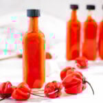 one rectangular bottle of hot sauce is surrounded by scotch bonnet peppers in the foreground, two additional bottles of hot sauce are out of focus behind it to the right