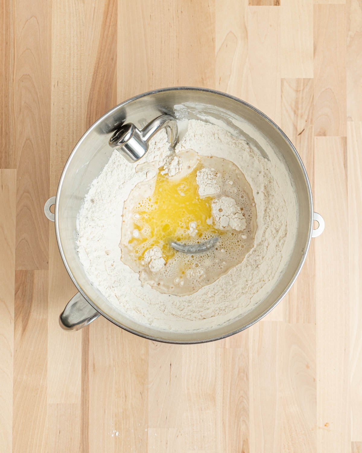 A stainless steel mixing bowl on a wooden surface contains flour, melted butter, and water, ready to be mixed for baking homemade pretzel bagels.