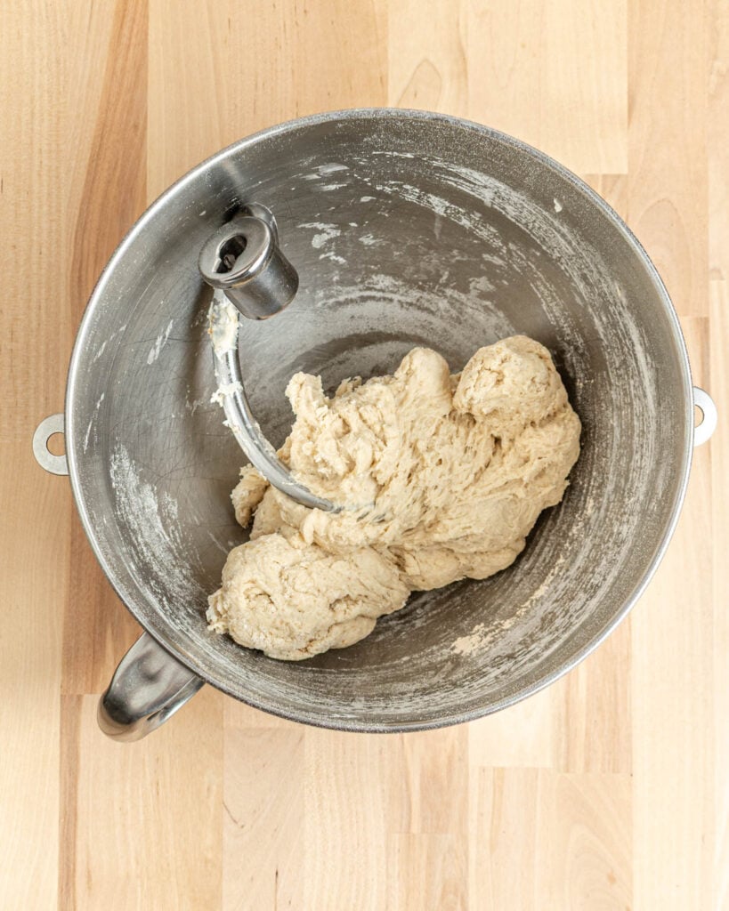 A metal mixing bowl with dough and a dough hook attachment sits on a light wooden surface, showing the process of kneading pretzel bagels dough.