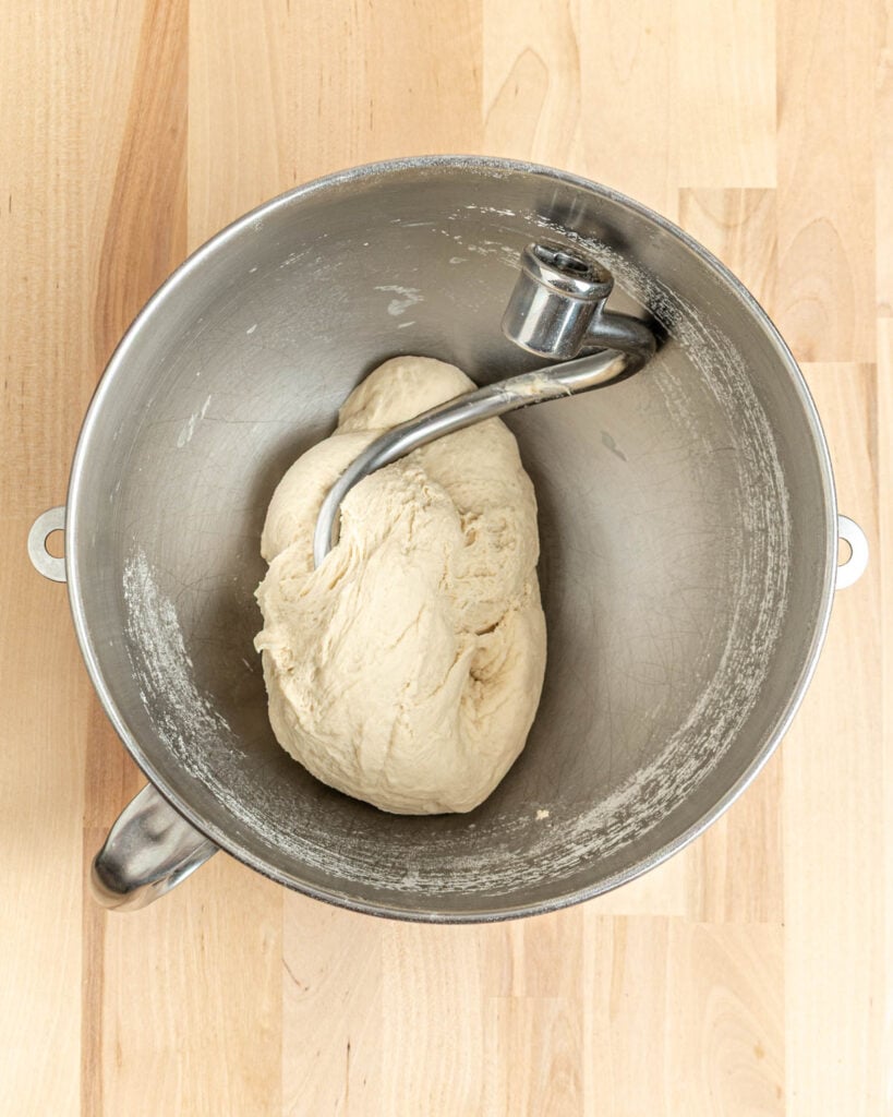 A ball of dough for pretzel bagels sits in a metal mixing bowl with a dough hook attachment on a light wooden surface.