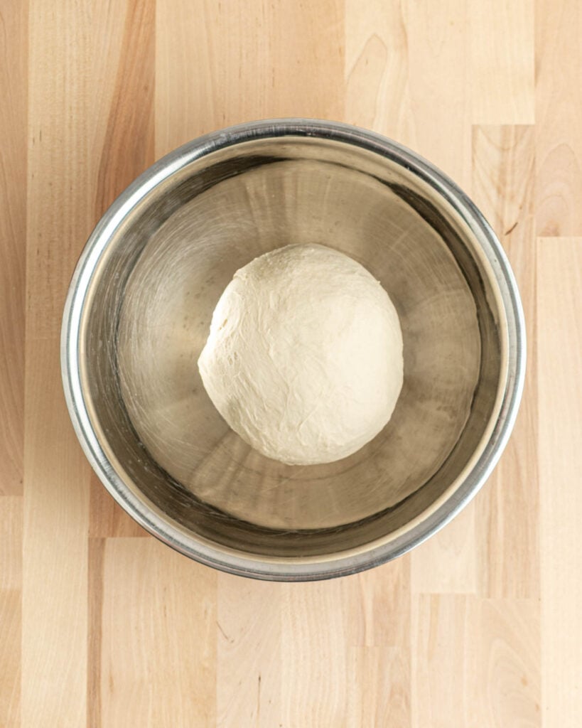 A tight ball of pretzel bagels dough rests in a large metal mixing bowl, placed on a light wooden surface.