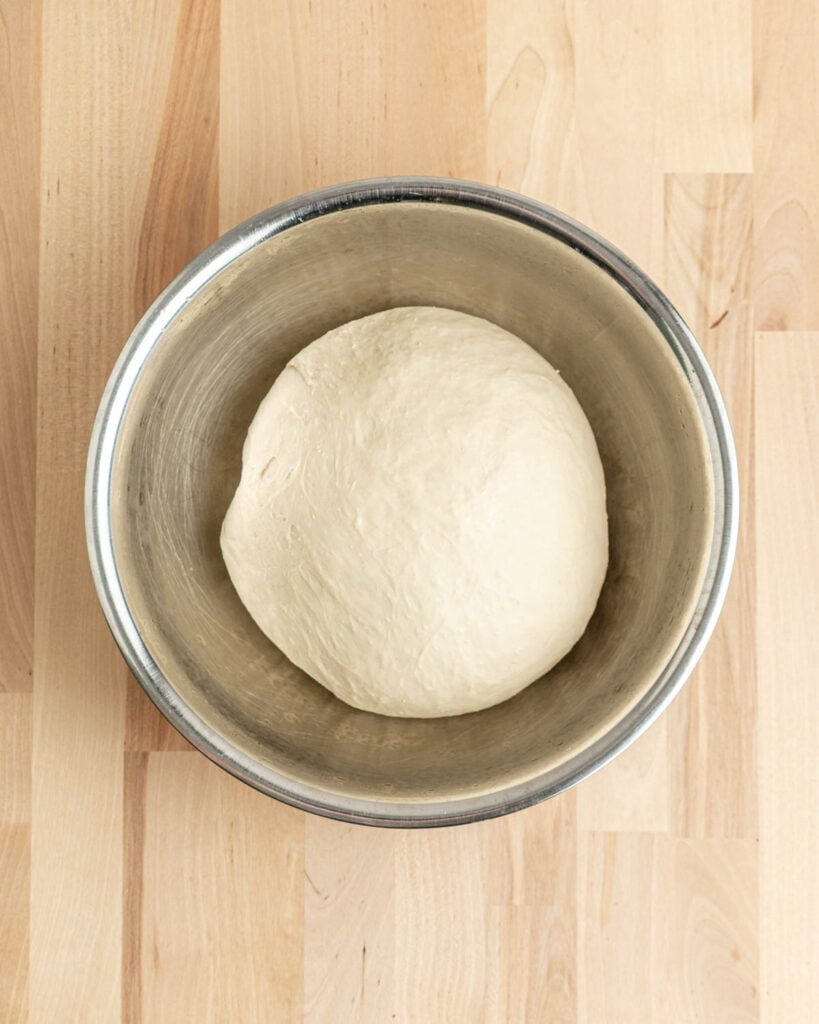 A round ball of dough, doubled in size, soon to become delicious pretzel bagels, rests in a metal mixing bowl on a light wooden surface.