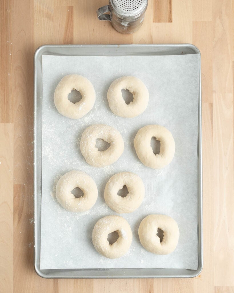 A baking sheet lined with parchment paper holds eight unbaked, floured pretzel bagels arranged in rows. A metal sifter sits above the tray on a light wooden surface.