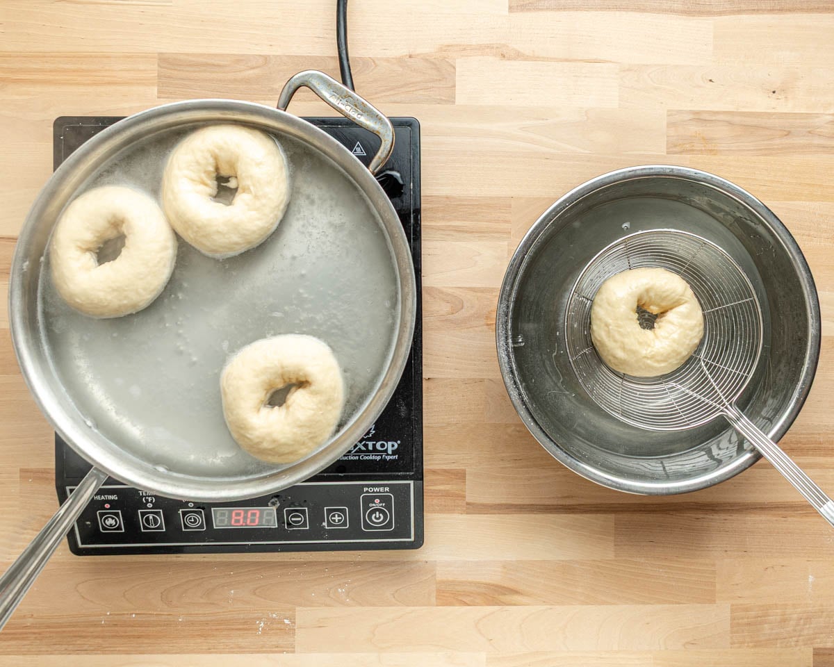 Overhead view of three pretzel bagels boiling in a pan on an electric stove and one bagel being dipped in the baking soda solution in a metal bowl with a wire spider, all on a wooden countertop.