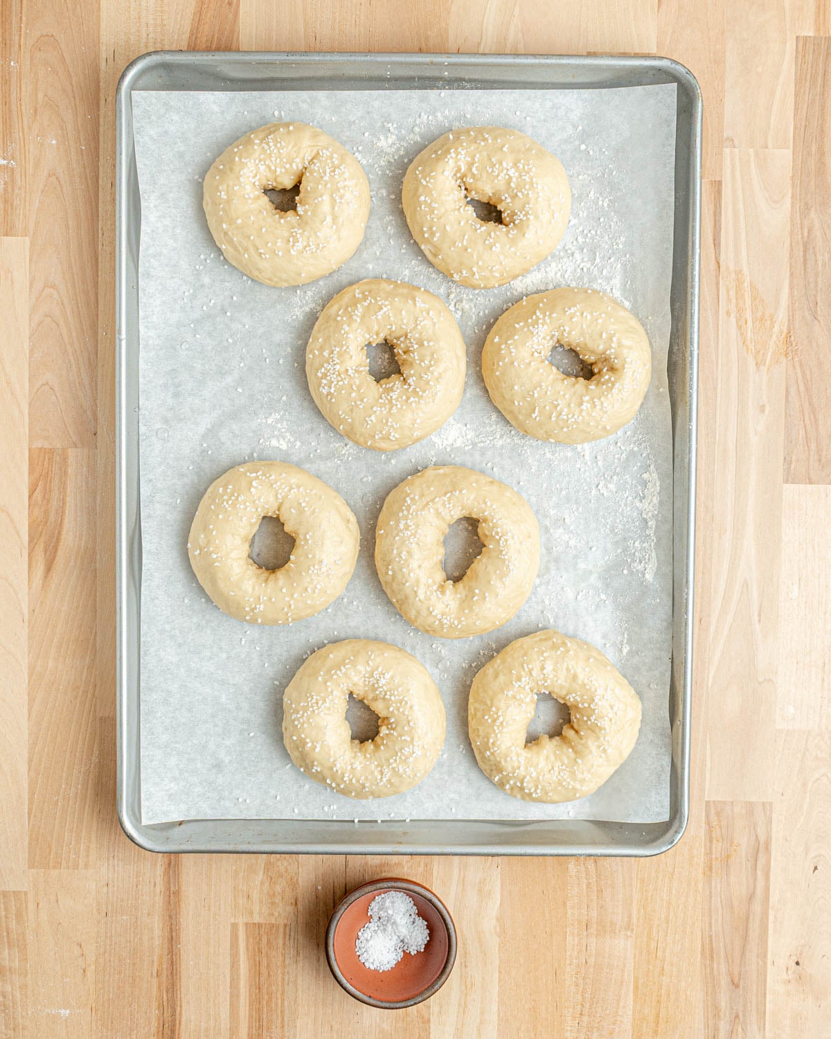 Eight unbaked pretzel bagels topped with coarse salt are arranged on a parchment-lined baking sheet. A small bowl of extra salt sits below the tray on a wooden surface.