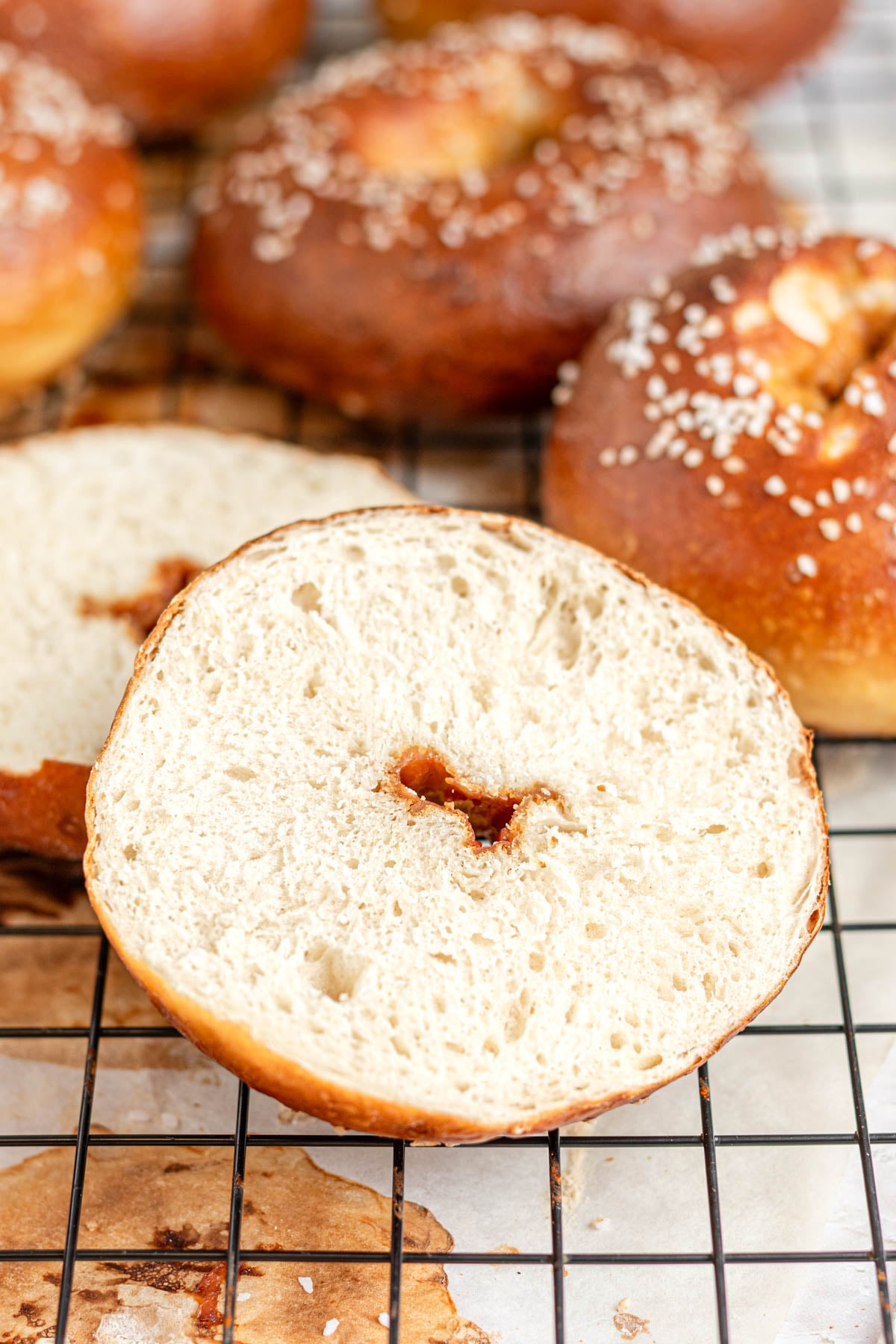 A sliced pretzel bagel with a light, airy interior rests on a cooling rack, surrounded by whole bagels topped with pretzel salt and a golden-brown crust.