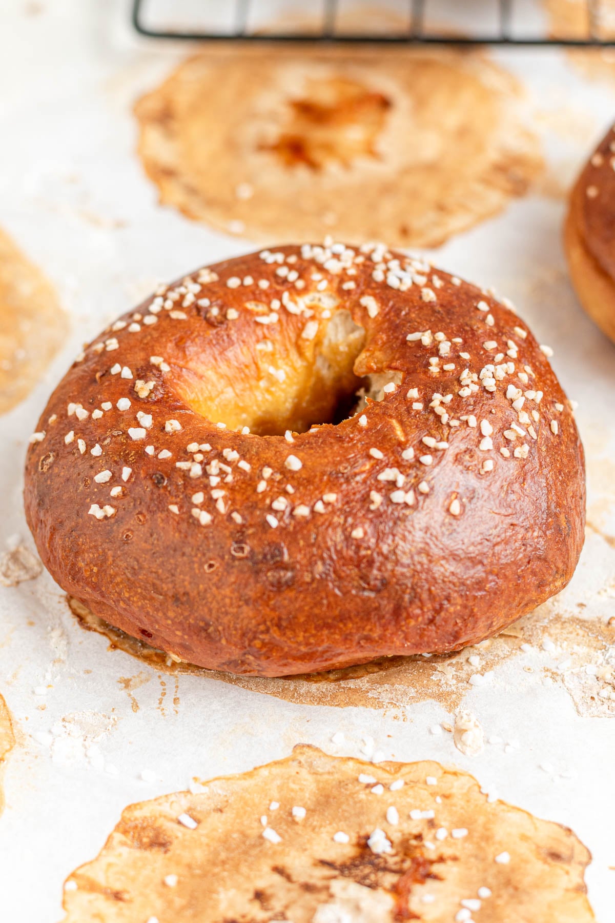 A golden-brown pretzel bagel topped with coarse salt sits on a sheet of parchment paper, surrounded by browned baking marks and crumbs.