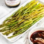 roasted asparagus on a rectangular serving platter. a small bowl of roasted garlic soy sauce is in front of it. a bottle of soy sauce is behind it.