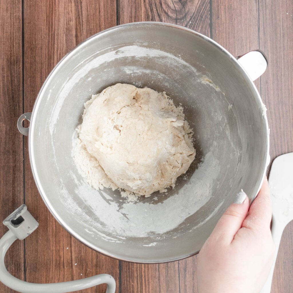 A hand holds a metal mixing bowl containing sourdough beer pretzel dough, surrounded by flour. The bowl sits on a wooden surface, with a dough hook and spatula nearby.