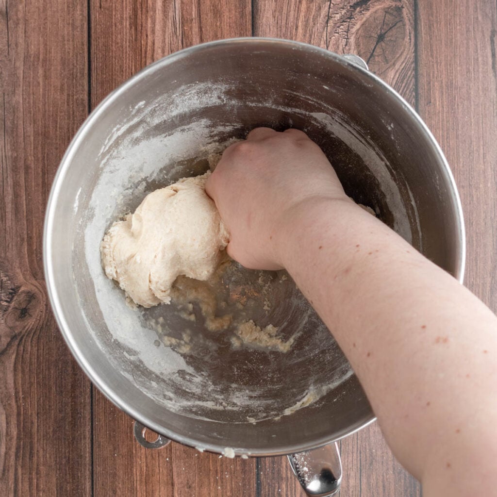 A person’s hand kneads dough for sourdough beer pretzels inside a metal mixing bowl, which sits on a brown wooden surface. Some flour remains visible along the bowl’s edges.