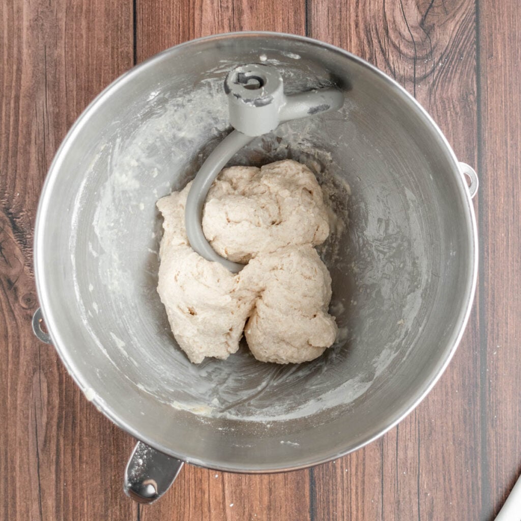 A metal mixing bowl with a dough hook attachment contains partially mixed sourdough beer pretzel dough. The bowl rests on a wooden surface.