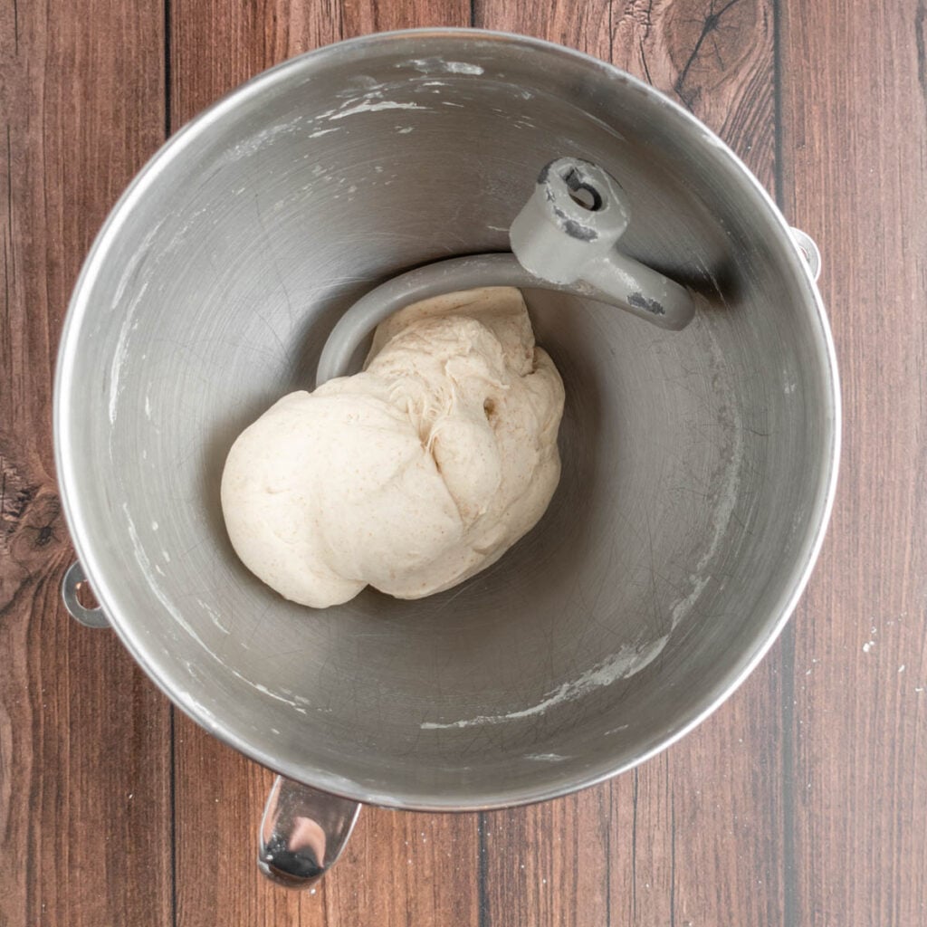 A ball of dough for sourdough beer pretzels sits in a metal mixing bowl with a dough hook attachment, placed on a wooden surface.