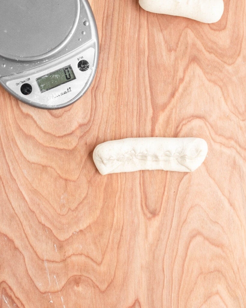 A digital kitchen scale with a zero reading sits on a wood surface next to two pieces of unbaked sourdough beer pretzel dough, one partly off-frame at the top and another in the center with visible creases.