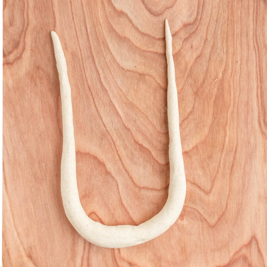 A piece of sourdough beer pretzel dough is rolled into a long rope shape and arranged in a U form on a wooden surface.