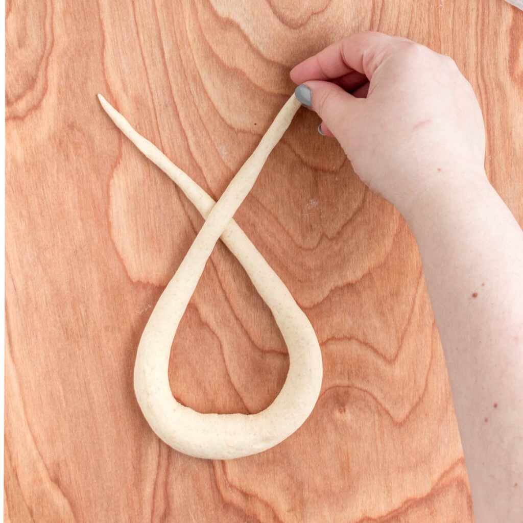 A hand shapes a long piece of sourdough beer pretzel dough into a loop with crossed ends on a wooden surface.