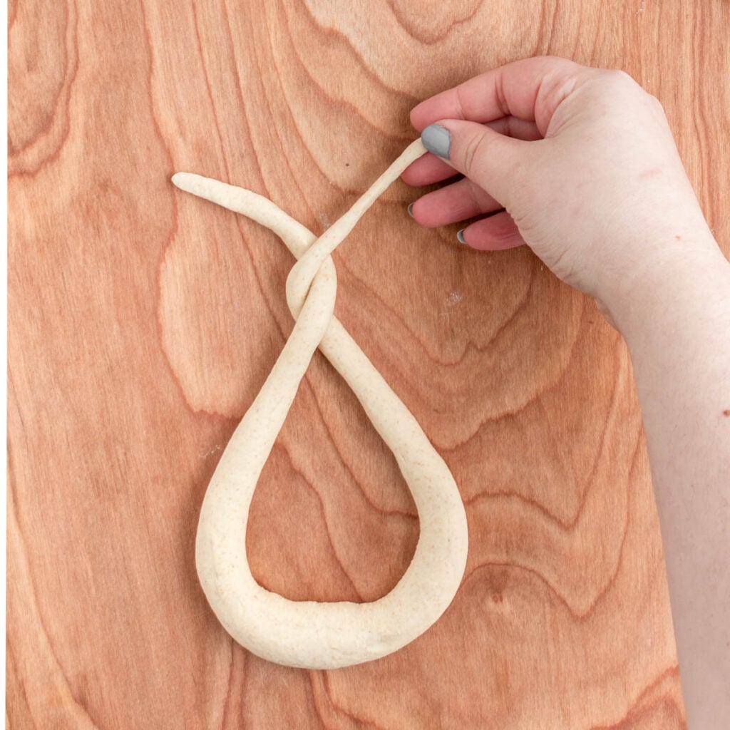 A hand with light blue nail polish twists the end of a rope of dough on a wooden surface, starting to shape it into sourdough beer pretzels.